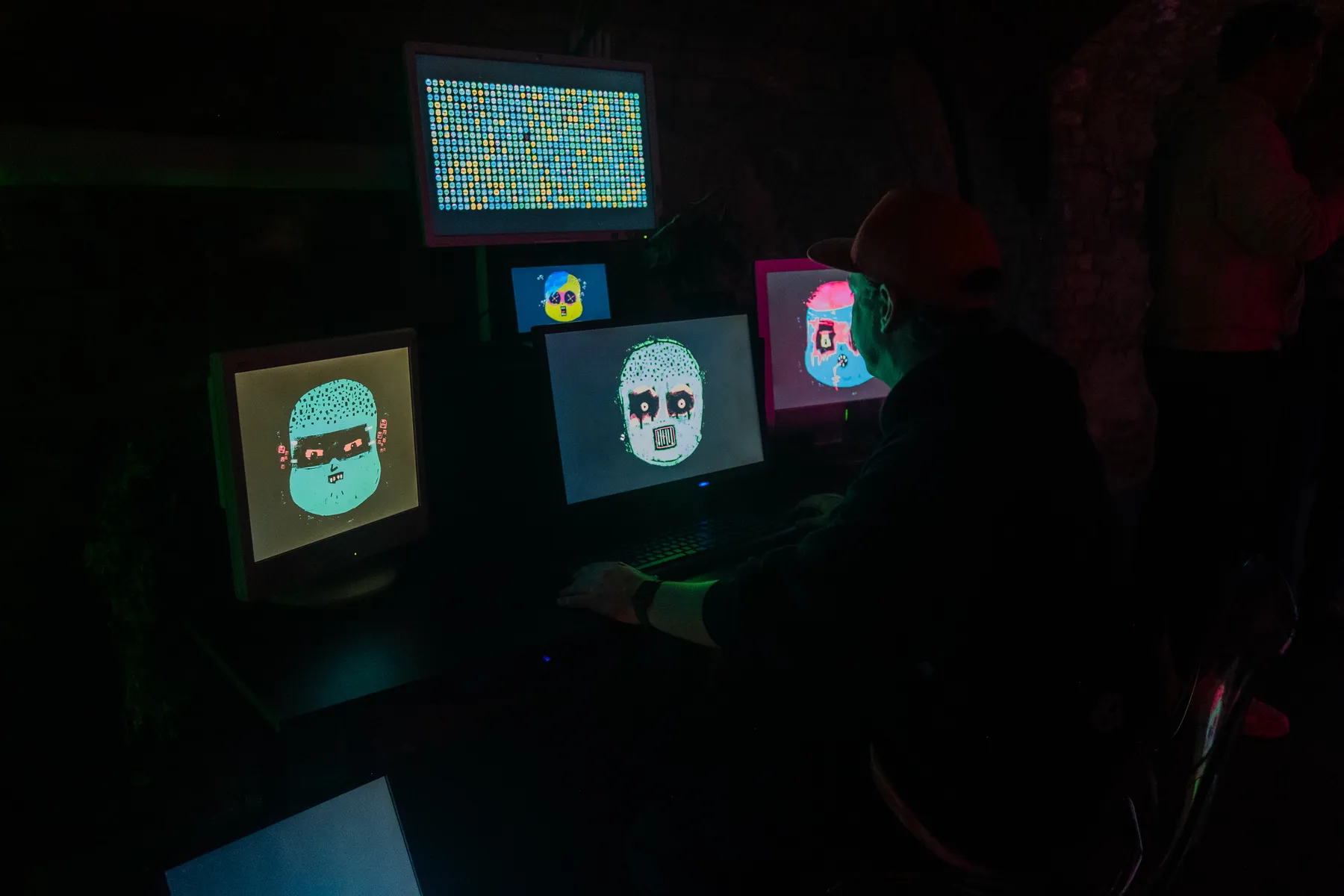 Abgedunkelter Raum, ein Sammler vor mehreren Bildschirmen, auf denen leuchtende Glitch-Porträts laufen