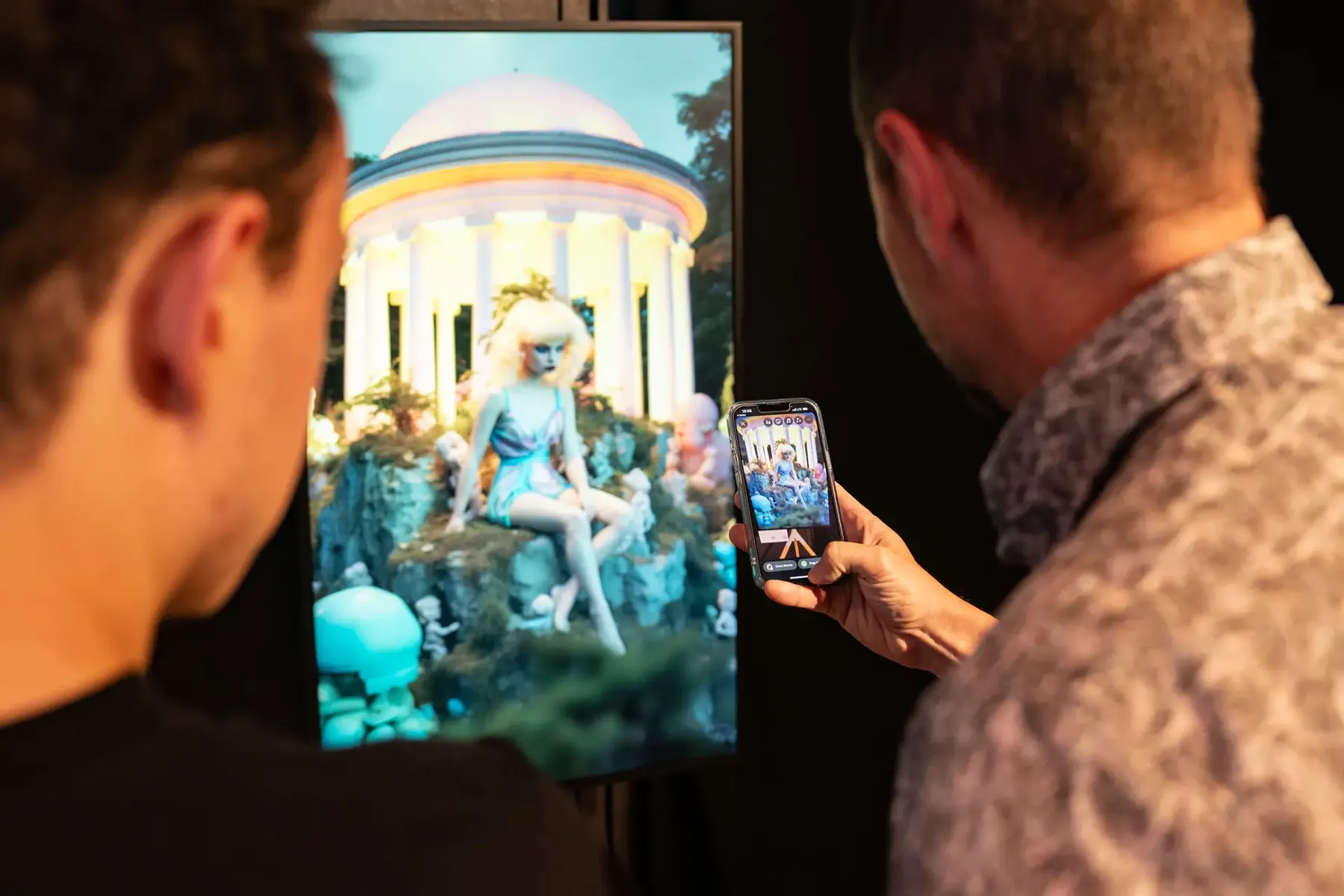 Zwei Betrachter vor einem gerahmten digitalen Werk auf einem Bildschirm, einer fotografiert es mit dem Telefon