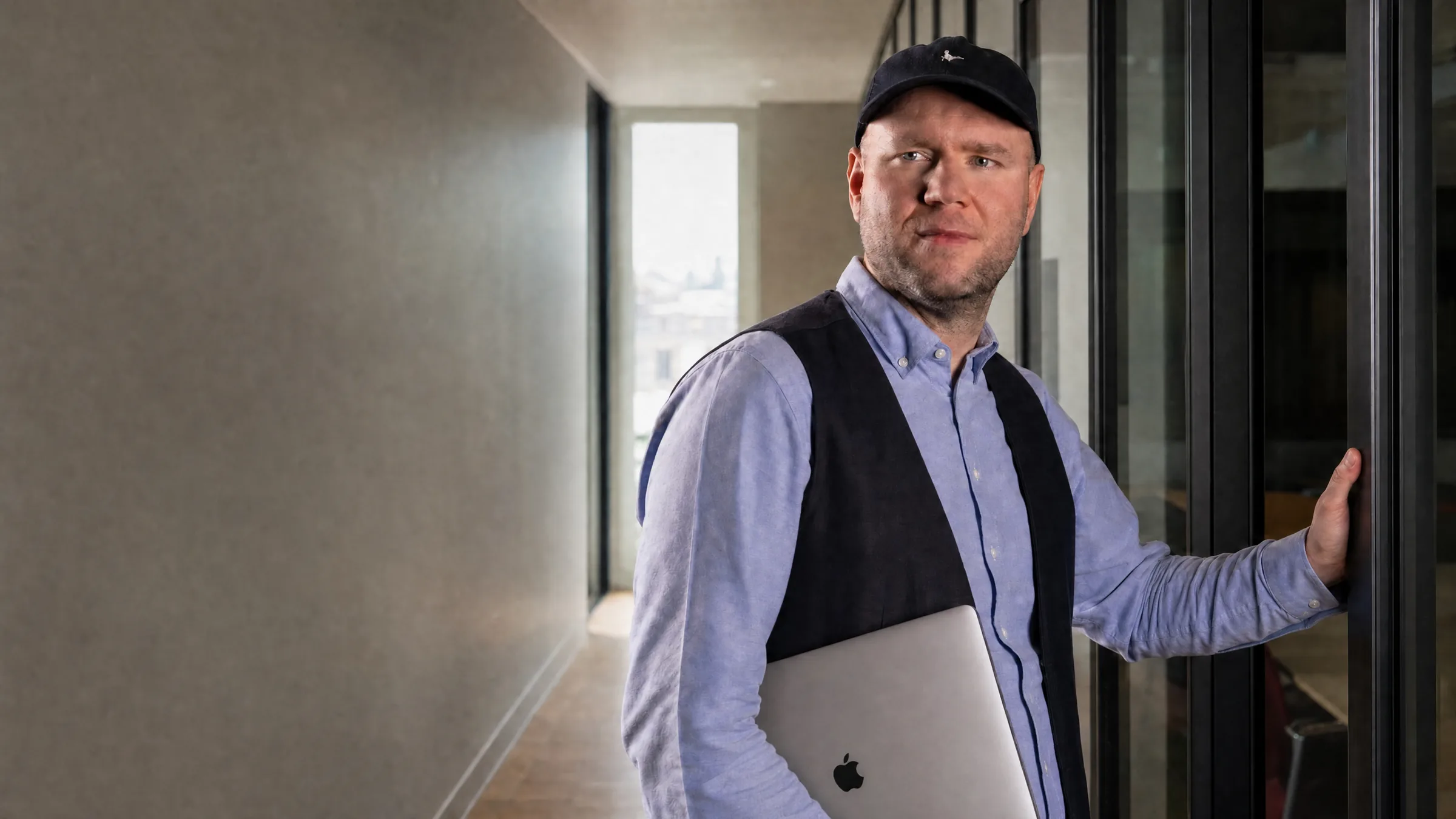 Mann mit Kappe und Weste steht im Bürogang an einer Glaswand, einen Laptop unter dem Arm